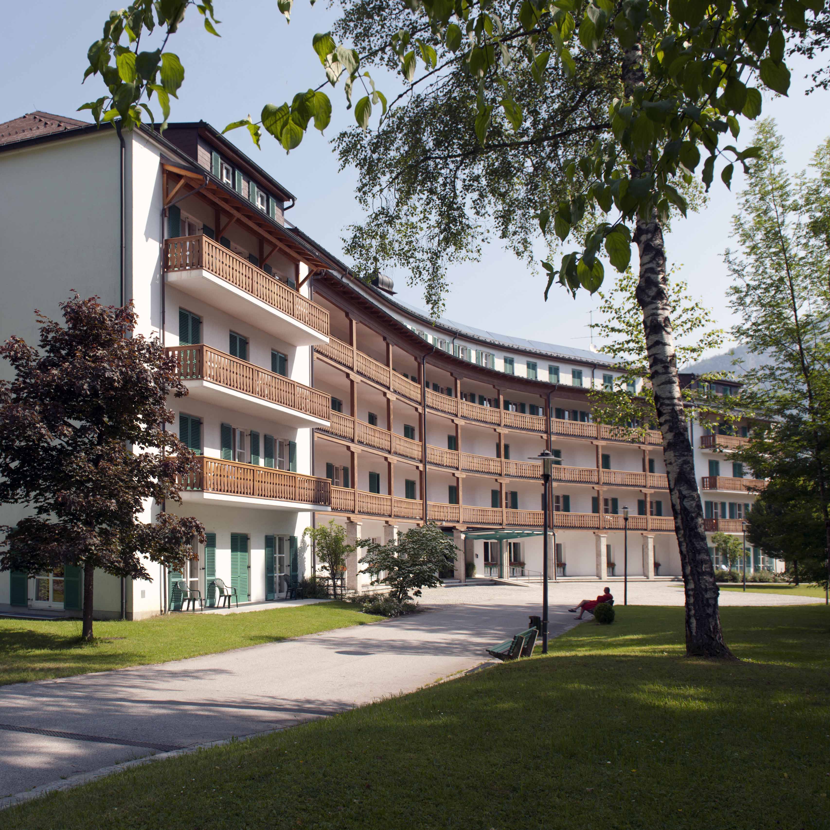 Außenansicht des Gesundheitszentrums Hanuschhof in Bad Goisern. Vor dem Haus ist eine Zufahrtsstraße zum Eingangsbereich. Recht daneben beginnt ein Teil des Parks mit Wiese, Bäumen und Bänken.